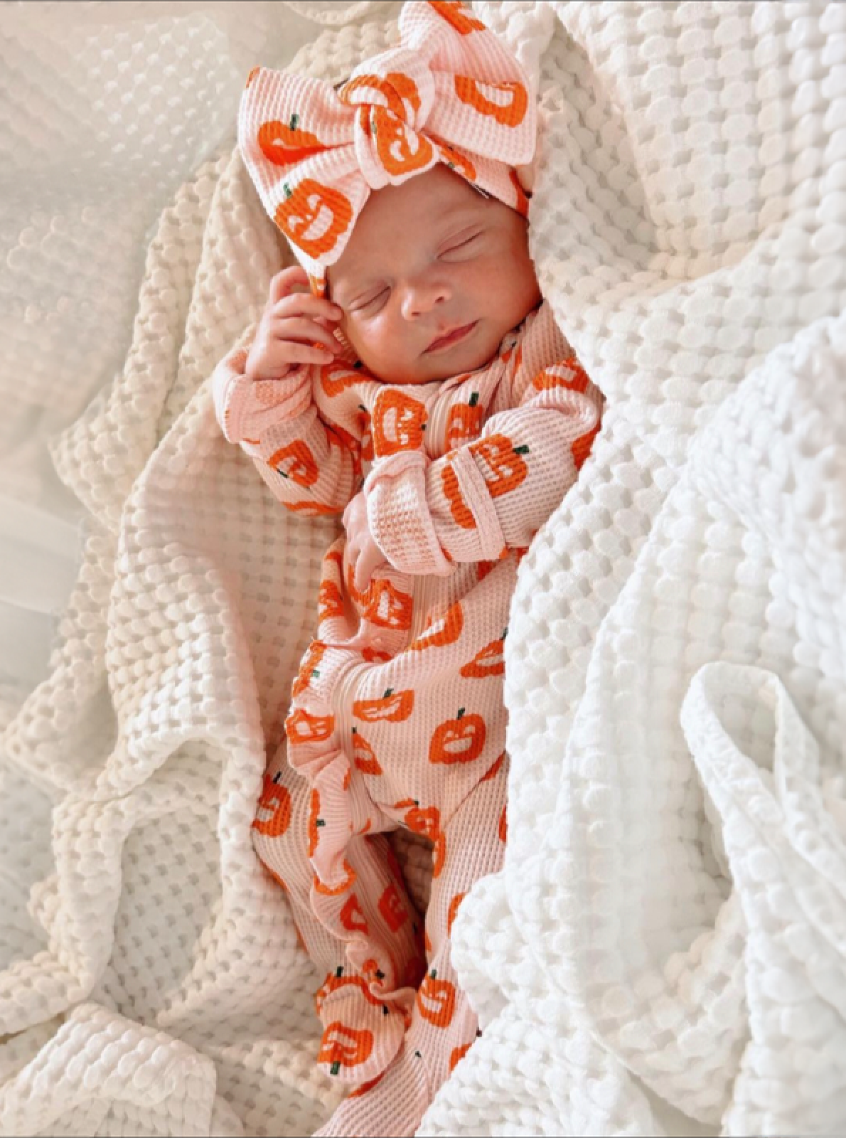 Sleeping baby in a pumpkin-patterned outfit and headband, nestled in white textured blankets.