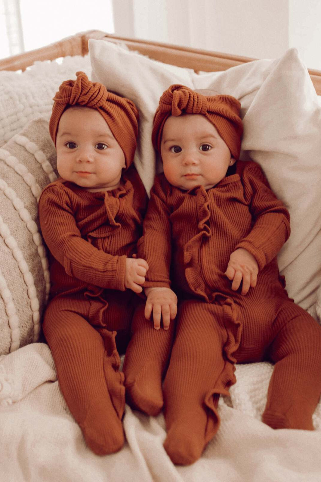 Two identical babies in brown outfits and headbands are sitting together on a cozy blanket.