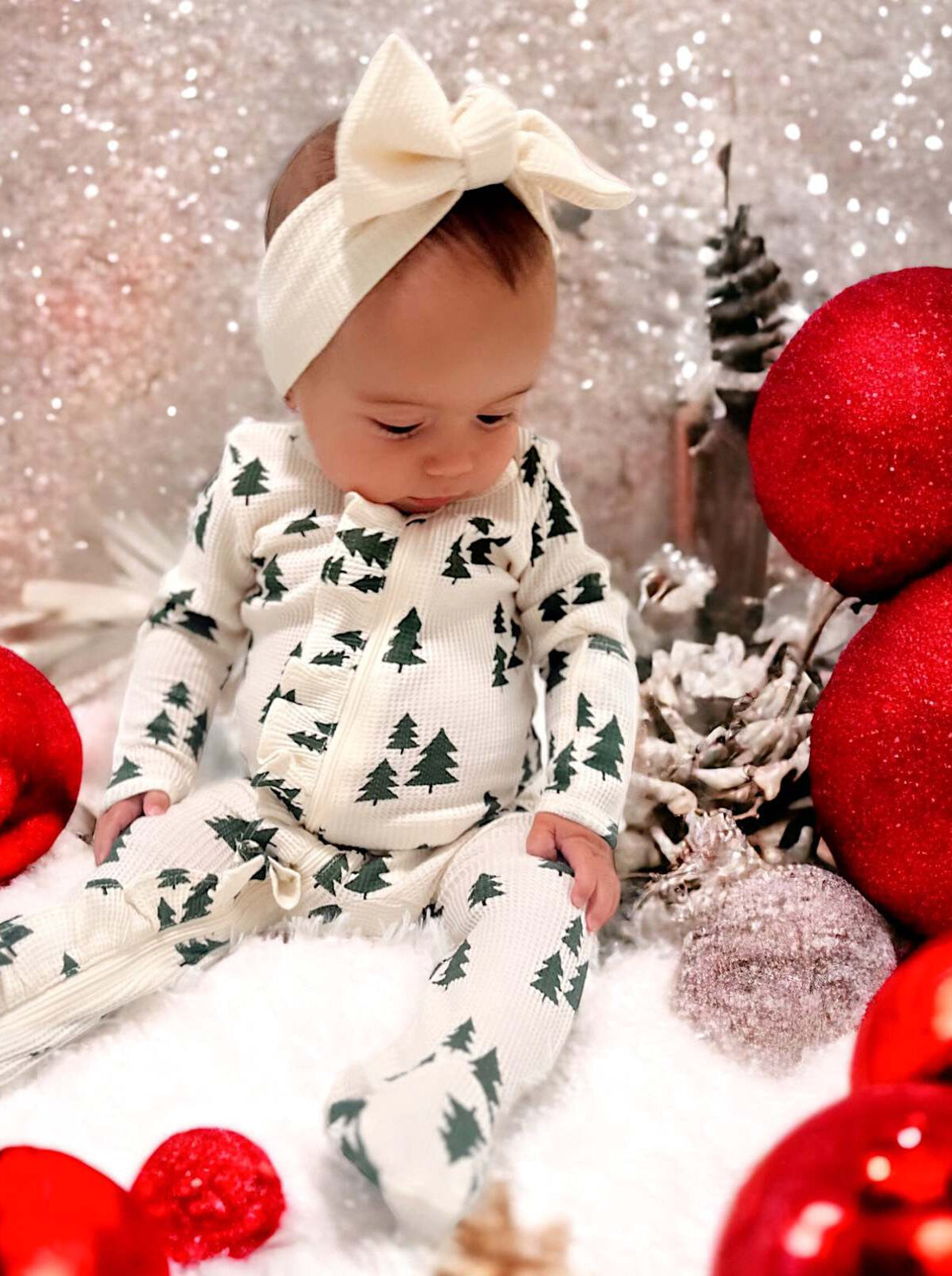 Baby in festive attire with tree patterns, surrounded by red ornaments and glittery decorations.