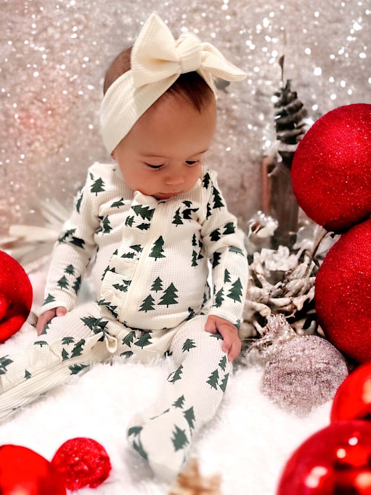 Baby in festive attire with tree patterns, surrounded by red ornaments and glittery decorations.