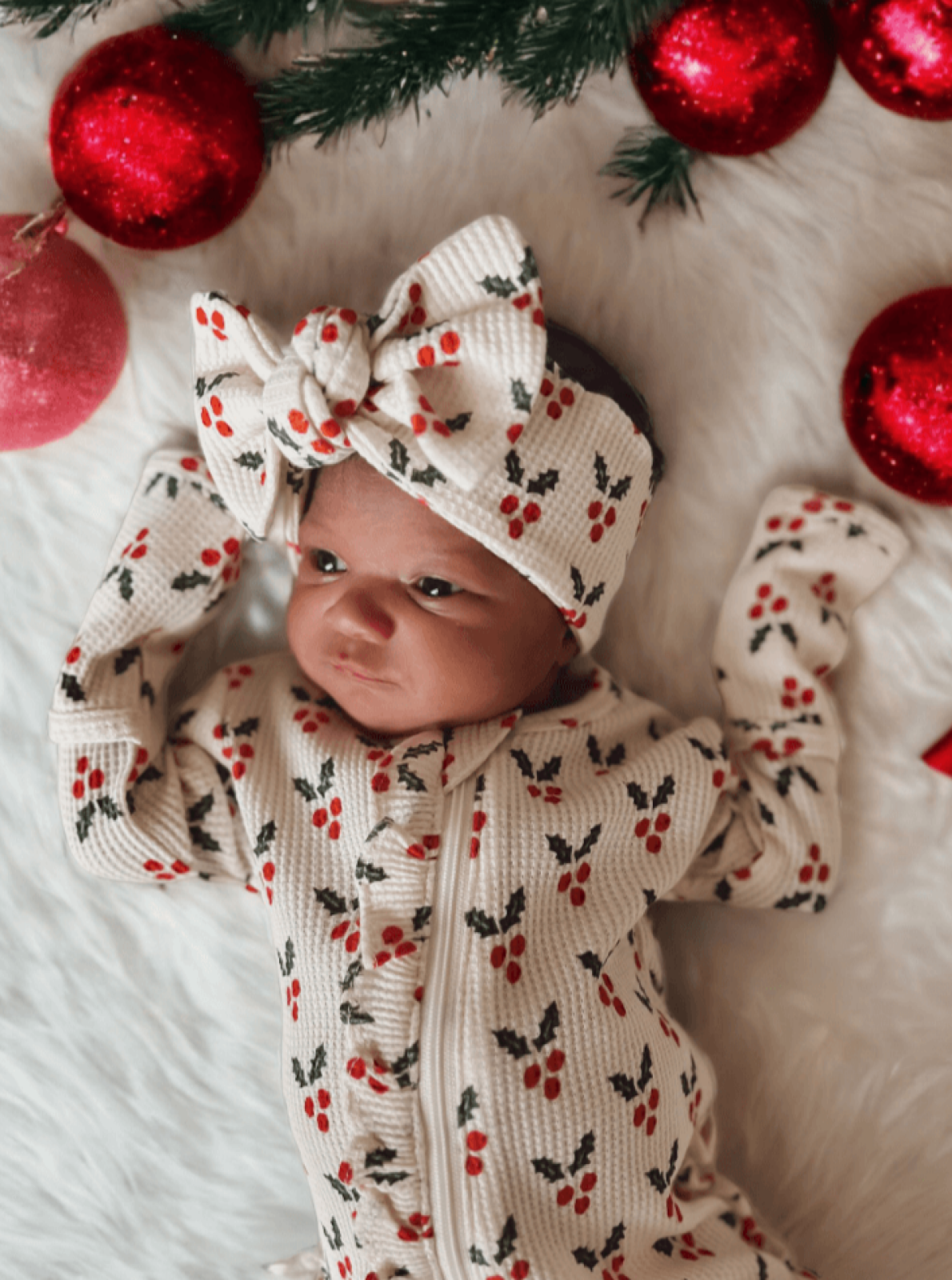 Newborn in a holiday outfit with red and green patterns, surrounded by red ornaments and festive decorations.