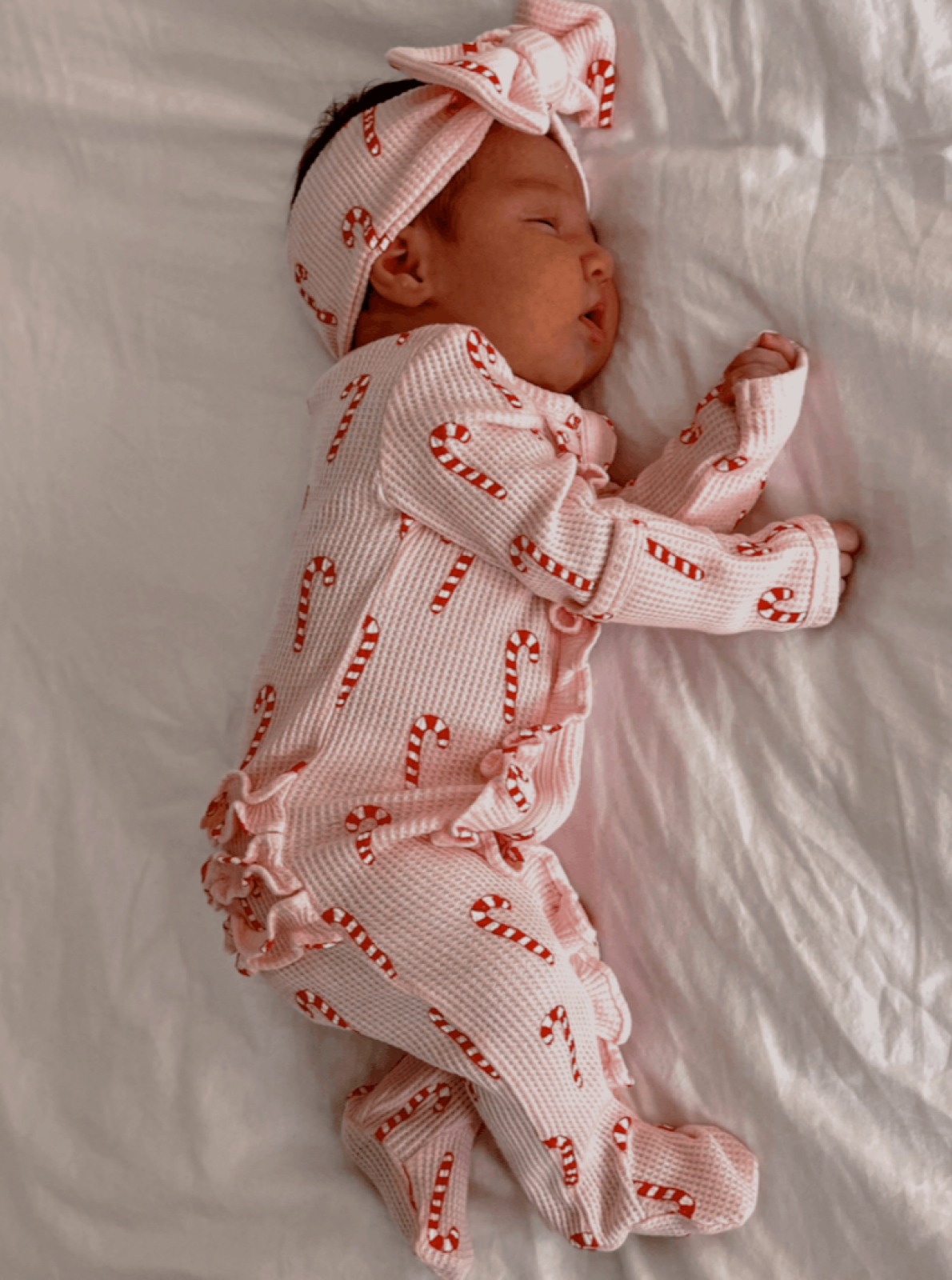 Sleeping baby in candy cane-patterned outfit and headband, resting on a white blanket.