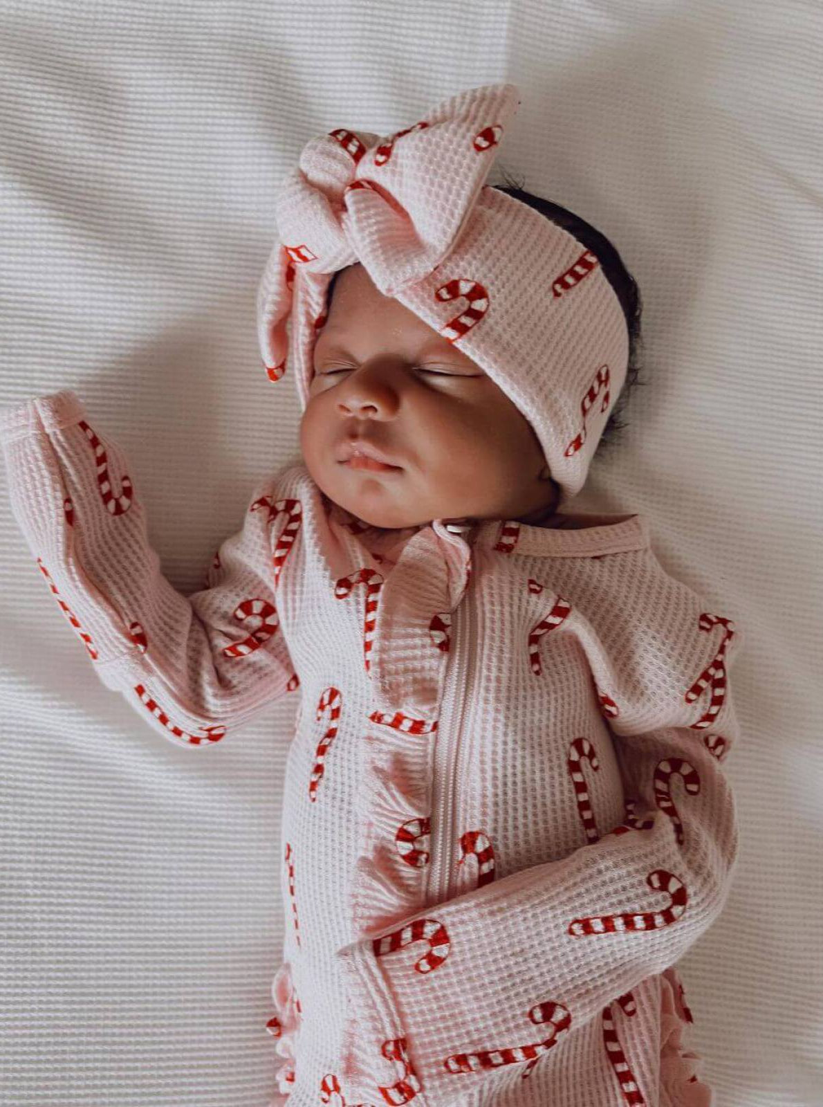 Baby girl asleep on a white blanket, dressed in pink festive outfit with candy cane patterns and a matching headband.