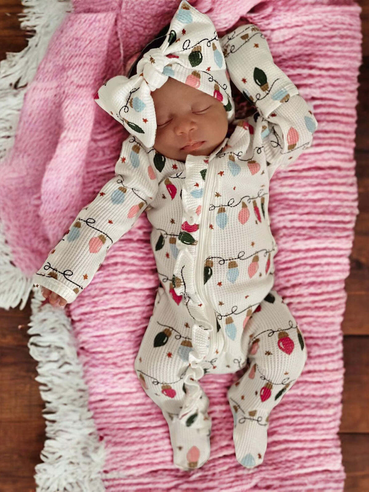 Newborn dressed in a light-colored outfit with Christmas lights pattern, lying on a pink textured blanket.