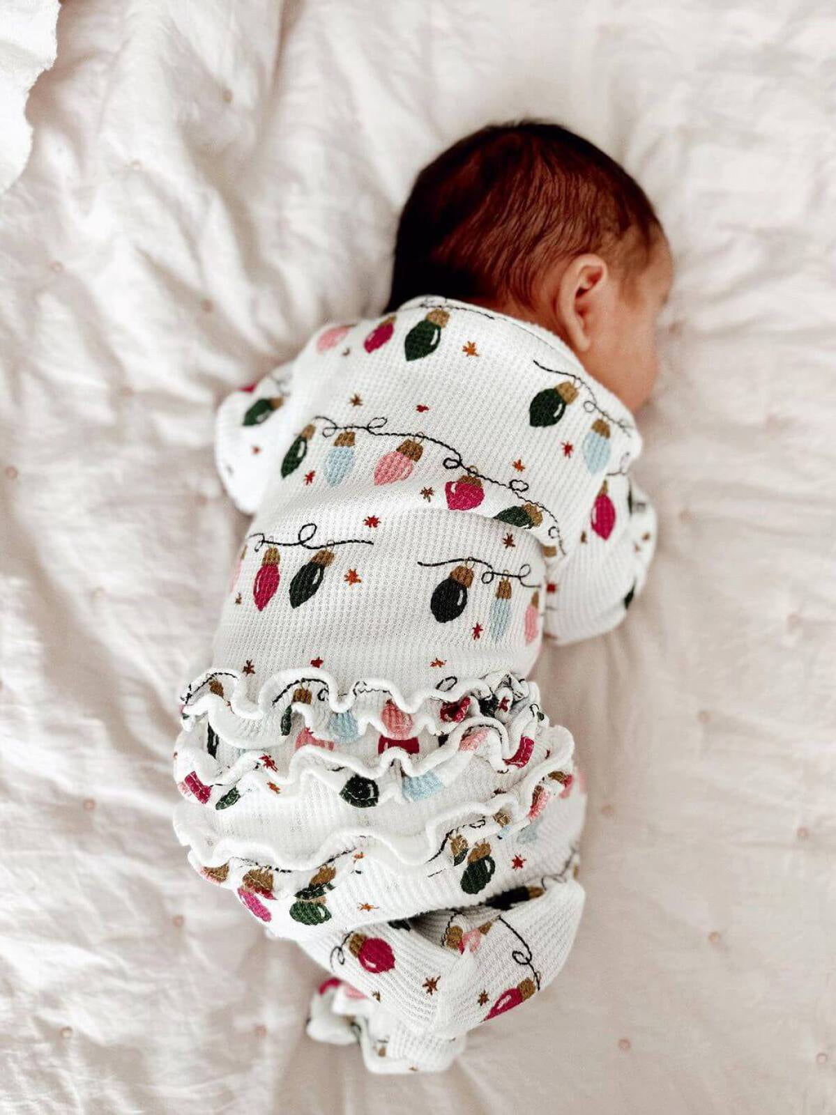 Infant sleeping on a light blanket, wearing a decorative onesie with holiday light patterns and ruffled detailing.