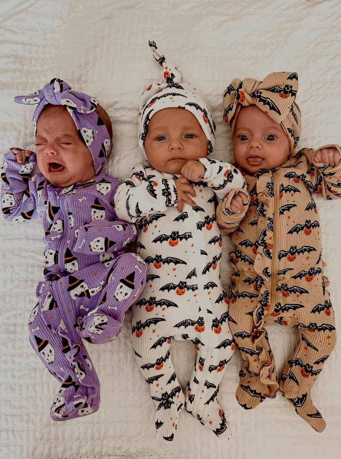 Three babies in Halloween-themed pajamas pose on a white blanket; one is crying, others are smiling.