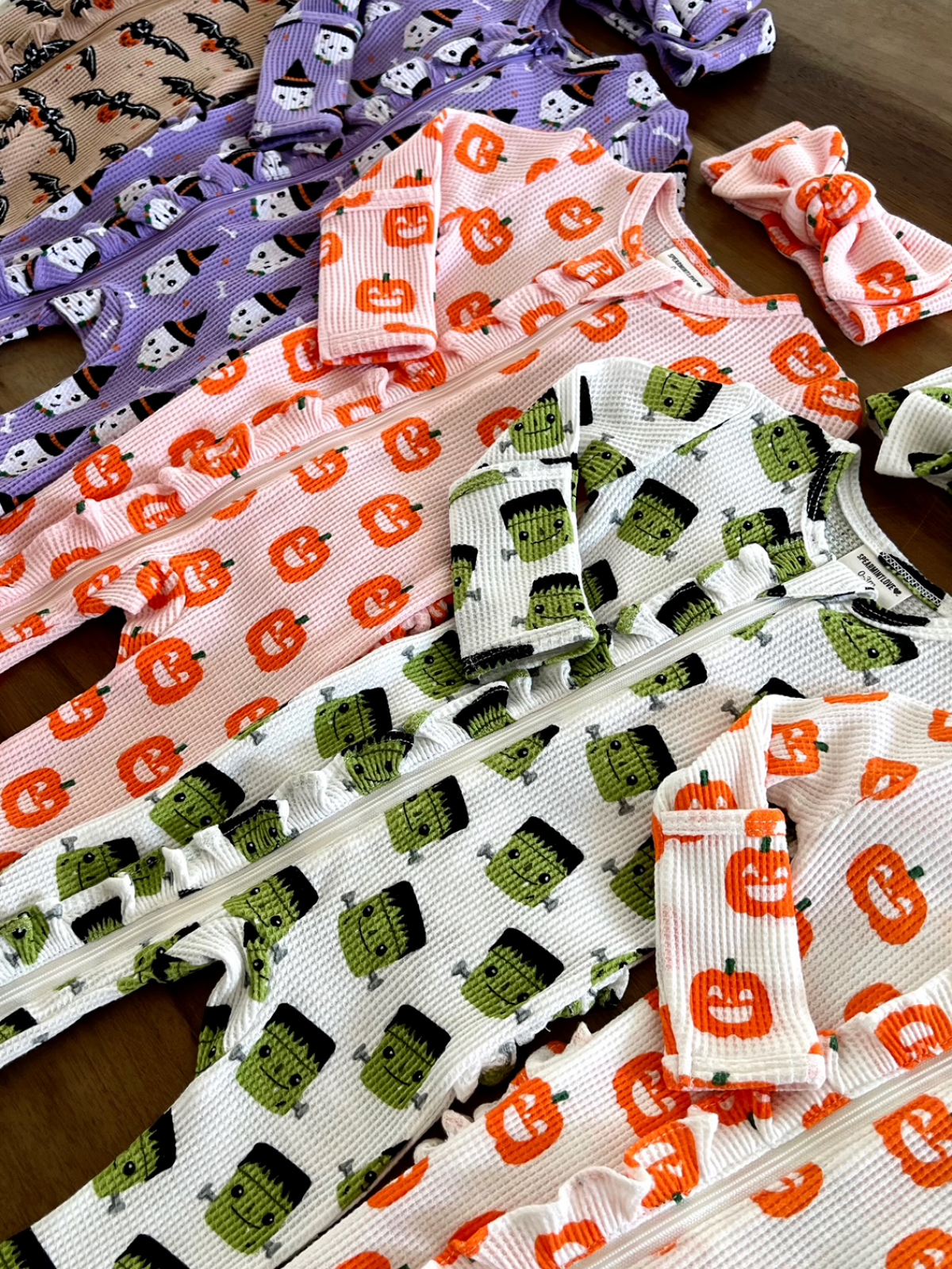 Colorful Halloween-themed baby outfits featuring pumpkins, bats, and Frankenstein designs on a wooden surface.