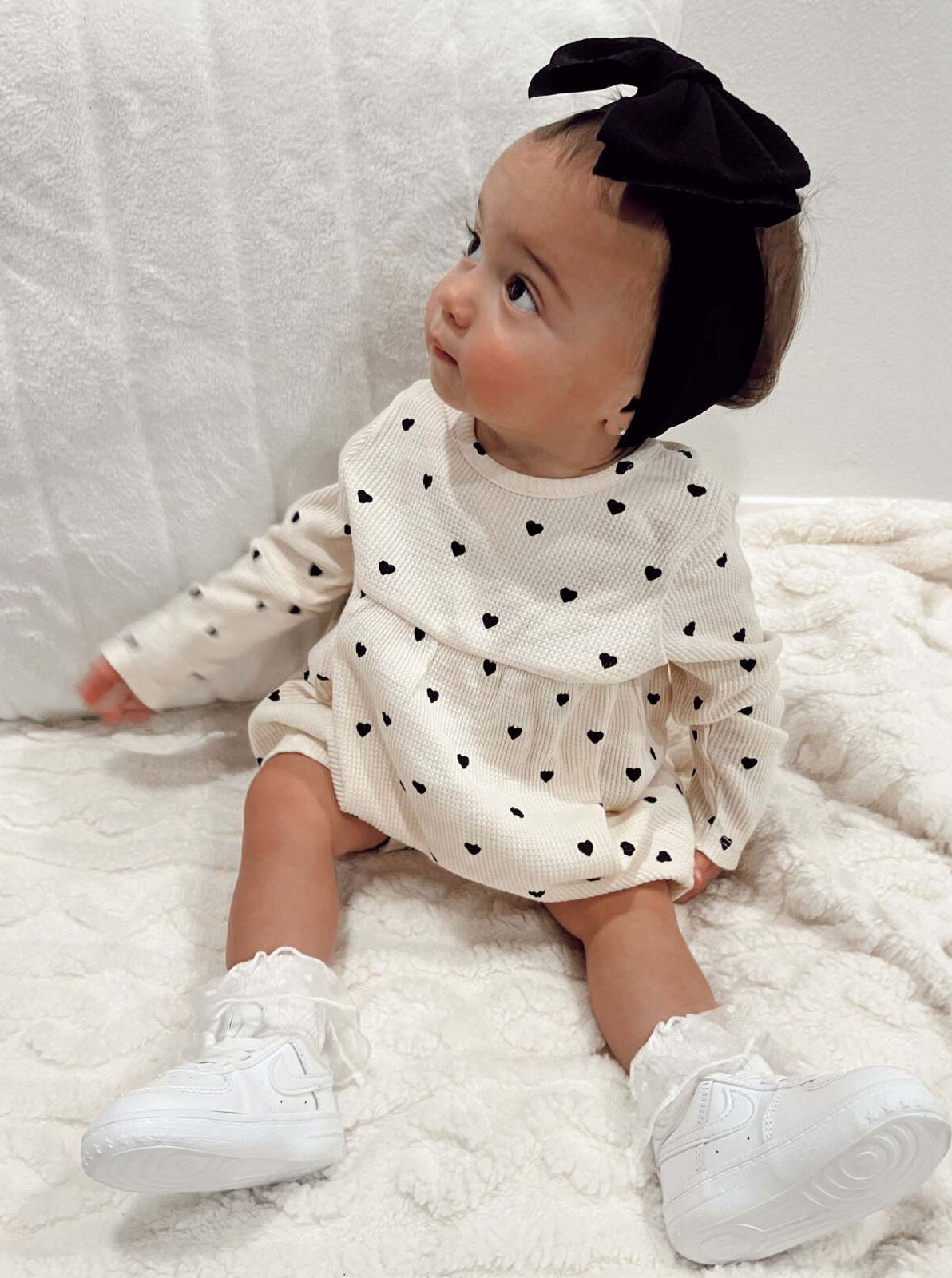 Baby girl with a black headband, wearing a heart-patterned dress, sitting on a cozy blanket.