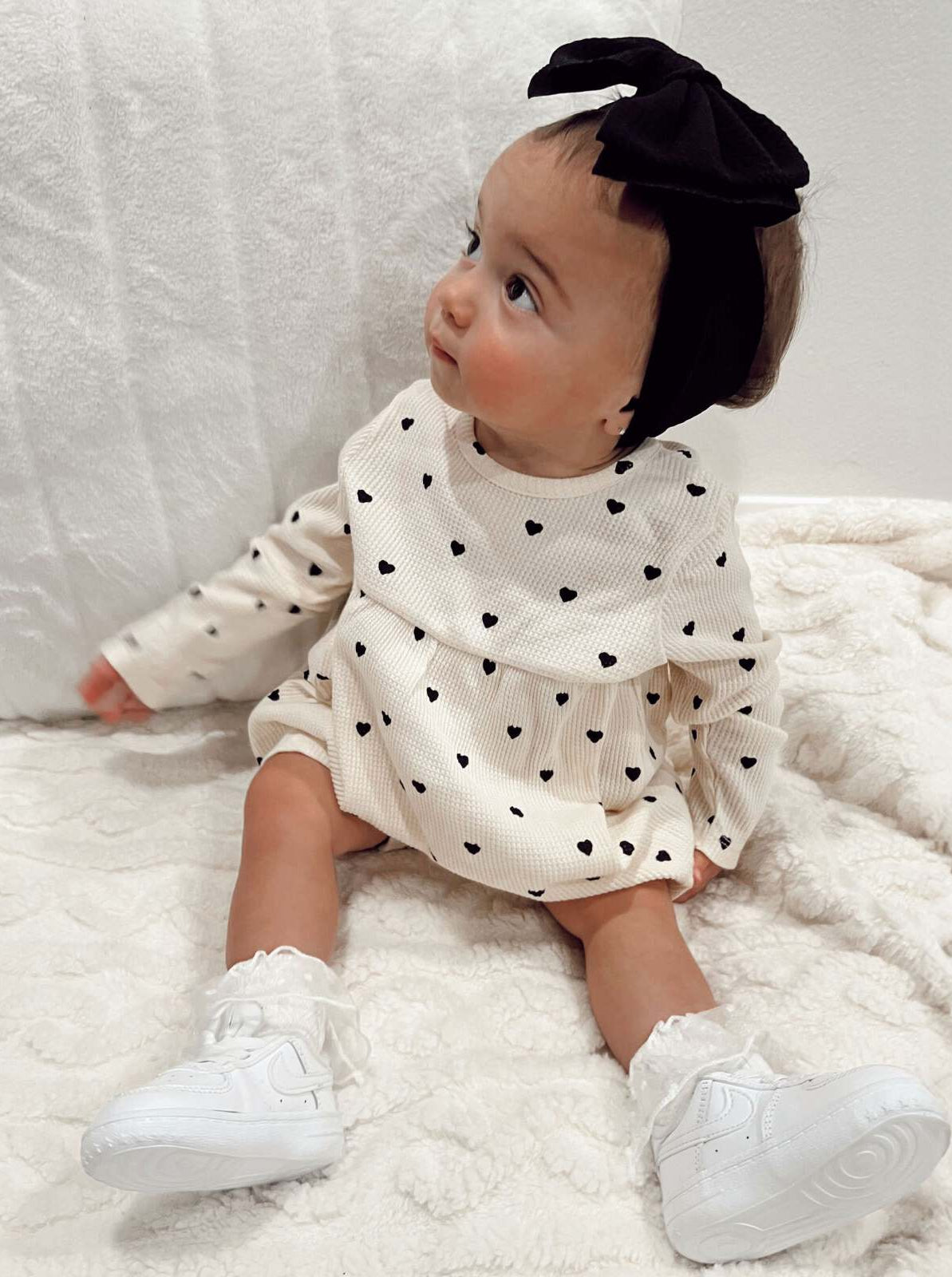 Baby girl with a black headband, wearing a heart-patterned dress, sitting on a cozy blanket.