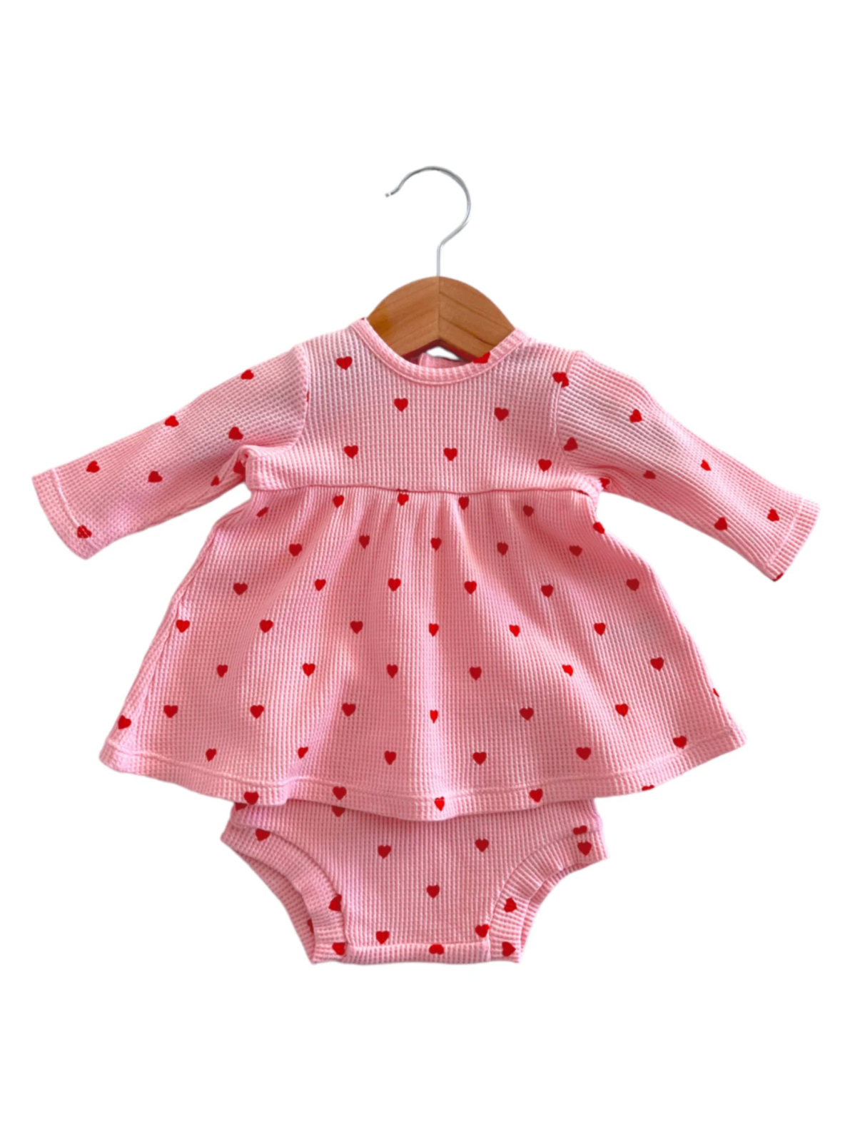Pink dress with red heart patterns, paired with matching bloomers, hanging on a wooden coat hanger.