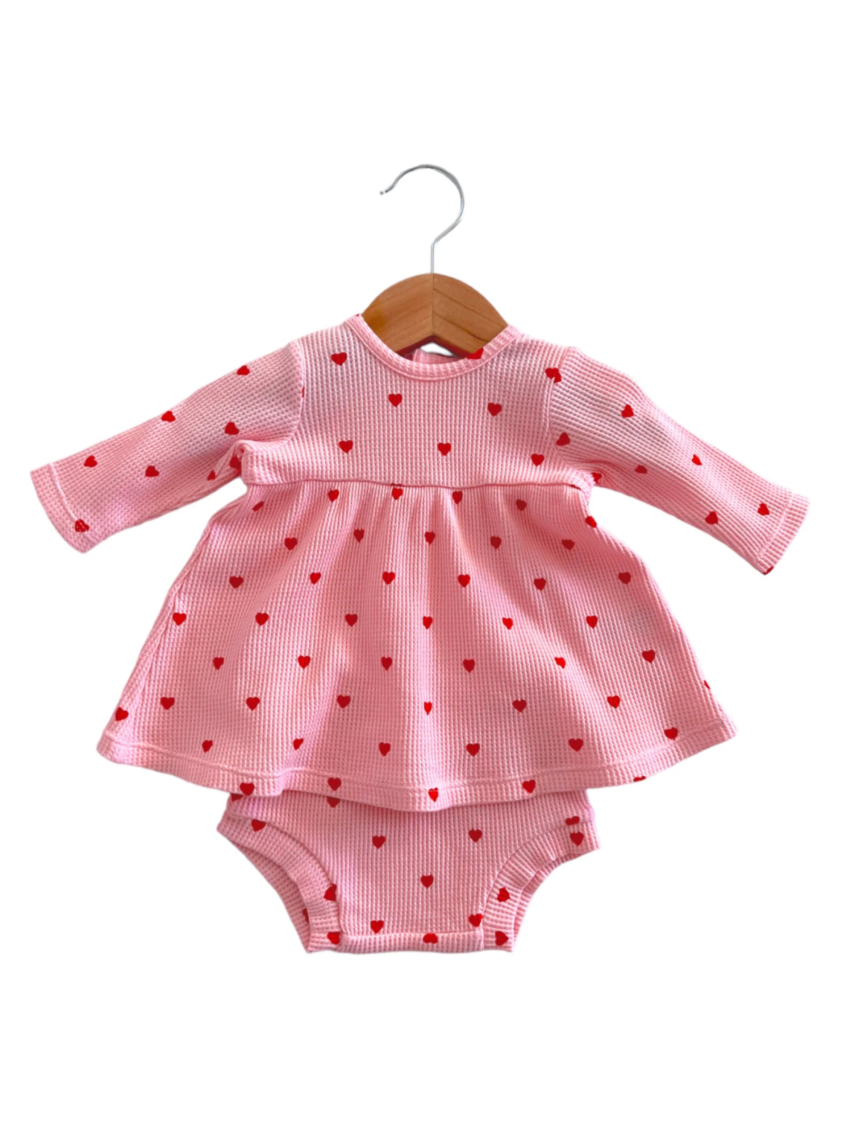 Pink dress with red heart patterns, paired with matching bloomers, hanging on a wooden coat hanger.