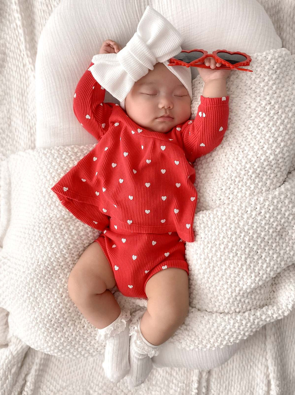 Baby girl in red outfit with heart patterns, wearing a large white bow, sleeps peacefully with red sunglasses nearby.