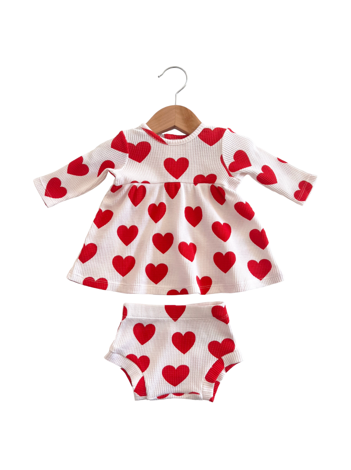 White baby dress with red heart patterns and matching shorts, hanging on a wooden hanger.