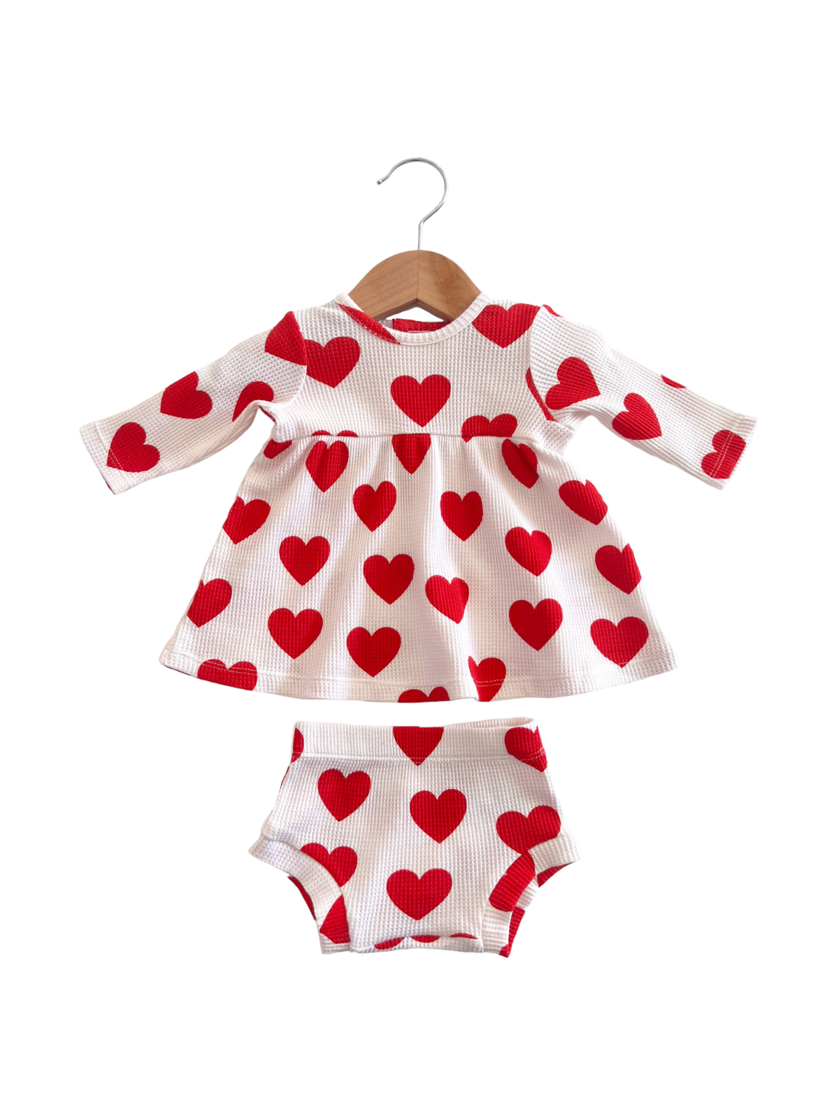White baby dress with red heart patterns and matching shorts, hanging on a wooden hanger.