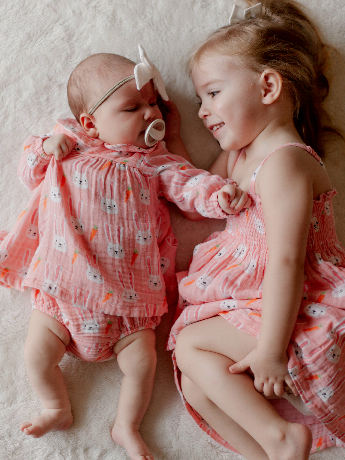 Two young girls in matching pink outfits with bunny prints, playing on a soft blanket. One is a baby, the other is older.