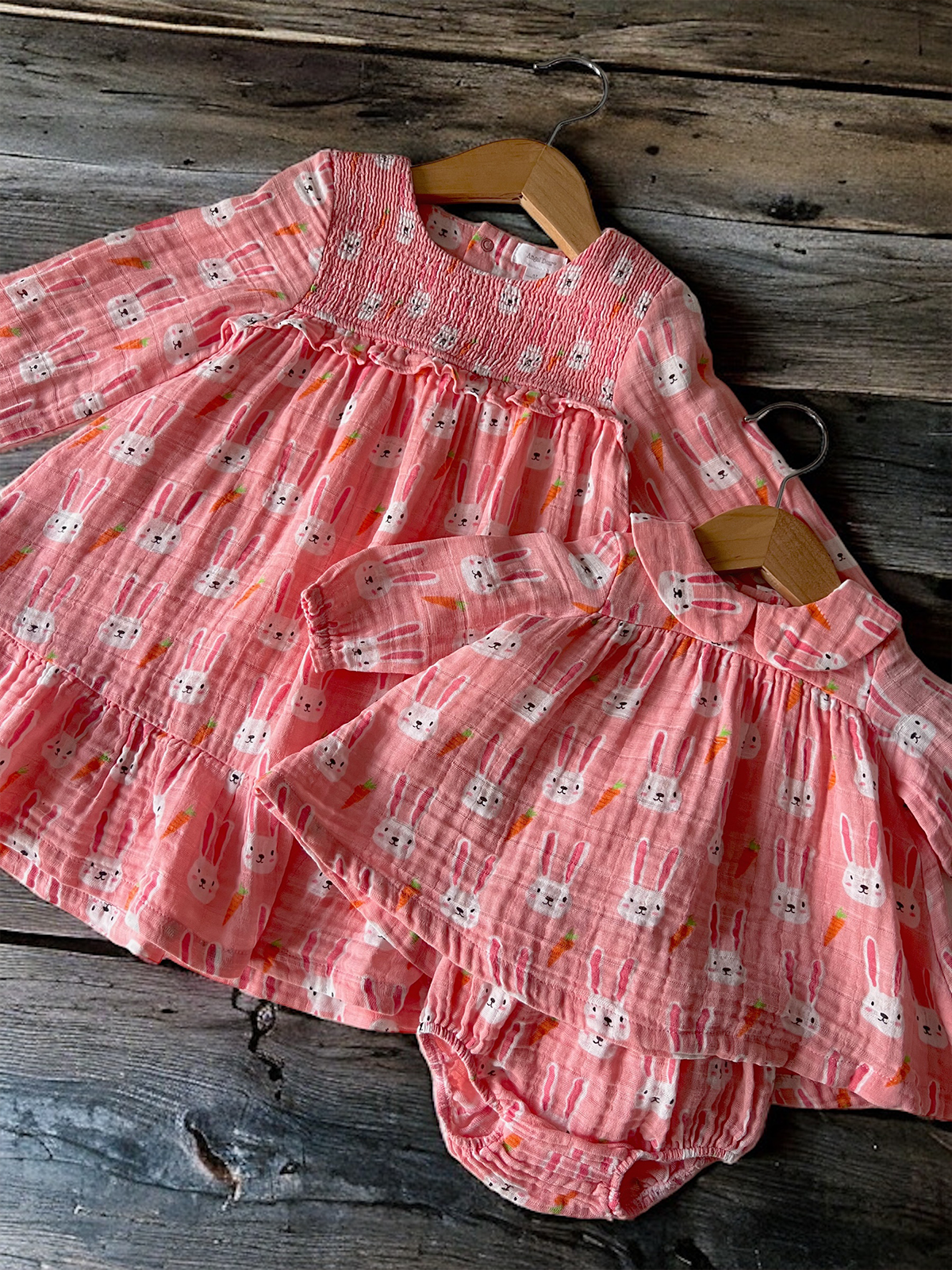 Two pink, bunny-patterned toddler dresses hanging on wooden hangers against a rustic wooden background.
