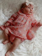 Baby in a pink bunny-print dress and bow, lying on a fluffy white blanket, gazing upward.