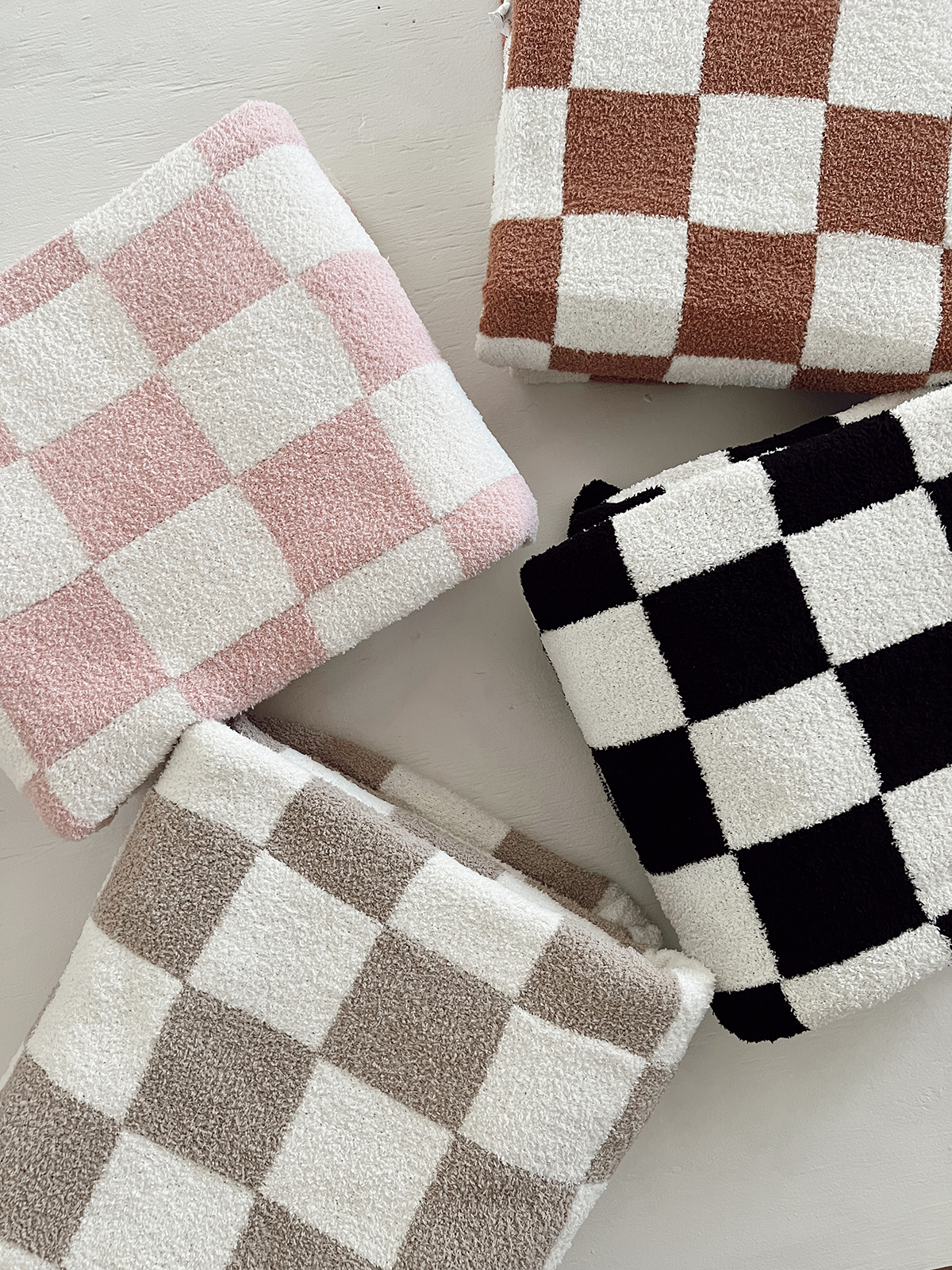 Four plush blankets with checkered patterns in pink, brown, white, and black, arranged on a light surface.