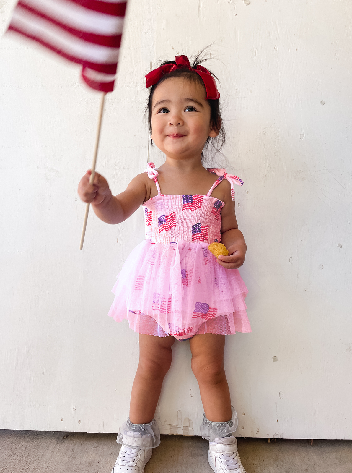 Smiling girl in patriotic dress holds a small American flag and a snack against a light background.