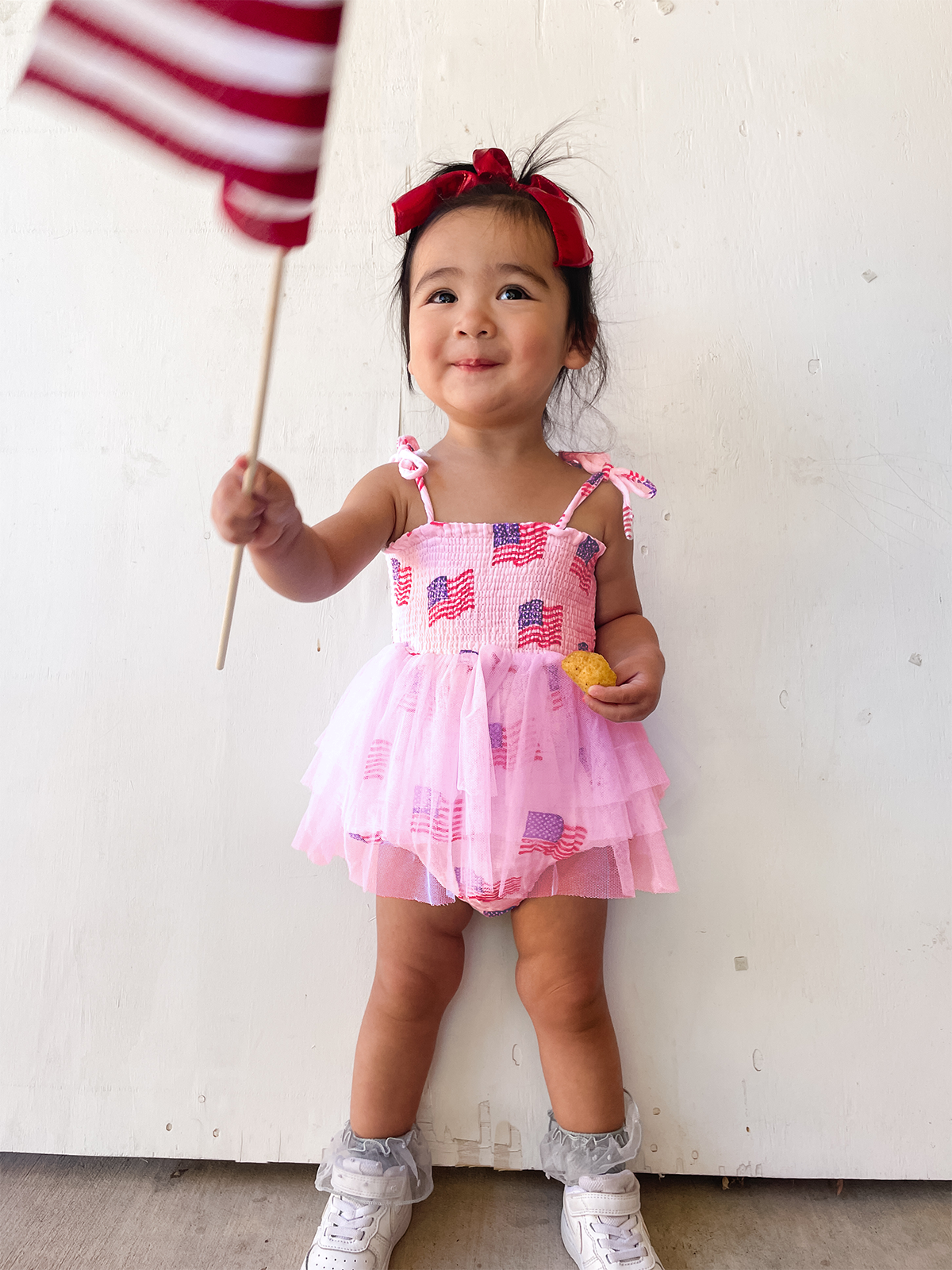 Smiling girl in patriotic dress holds a small American flag and a snack against a light background.