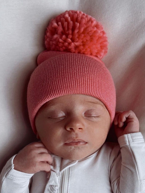 Sleeping baby wearing a pink beanie with a pom-pom on a soft white surface.