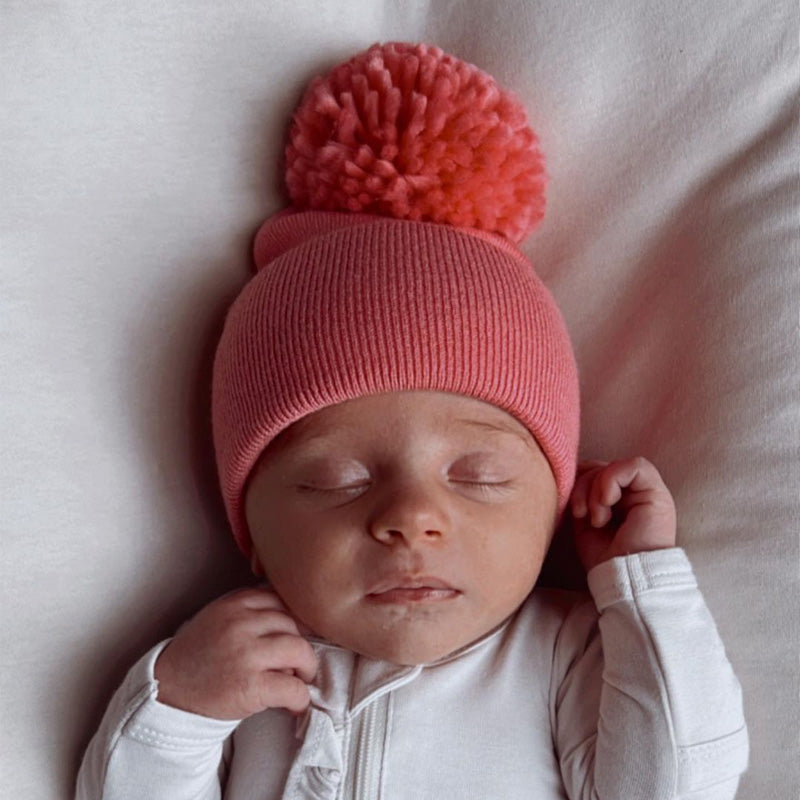 Sleeping baby wearing a pink beanie with a pom-pom on a soft white surface.