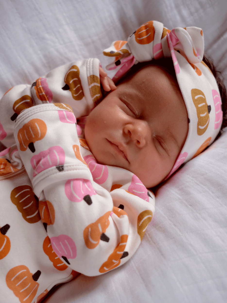 Baby girl sleeping peacefully in a pumpkin-patterned outfit and headband on a white blanket.