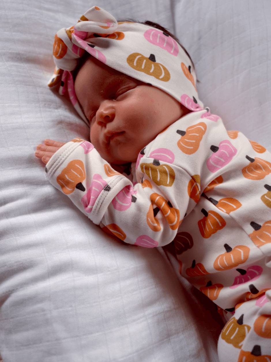 Baby sleeping peacefully on a white pillow, wearing a pumpkin-patterned onesie and matching headband.