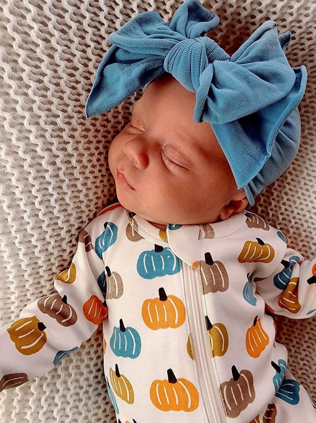 Sleeping baby in a pumpkin-patterned onesie, wearing a large blue bow headband, resting on a textured blanket.