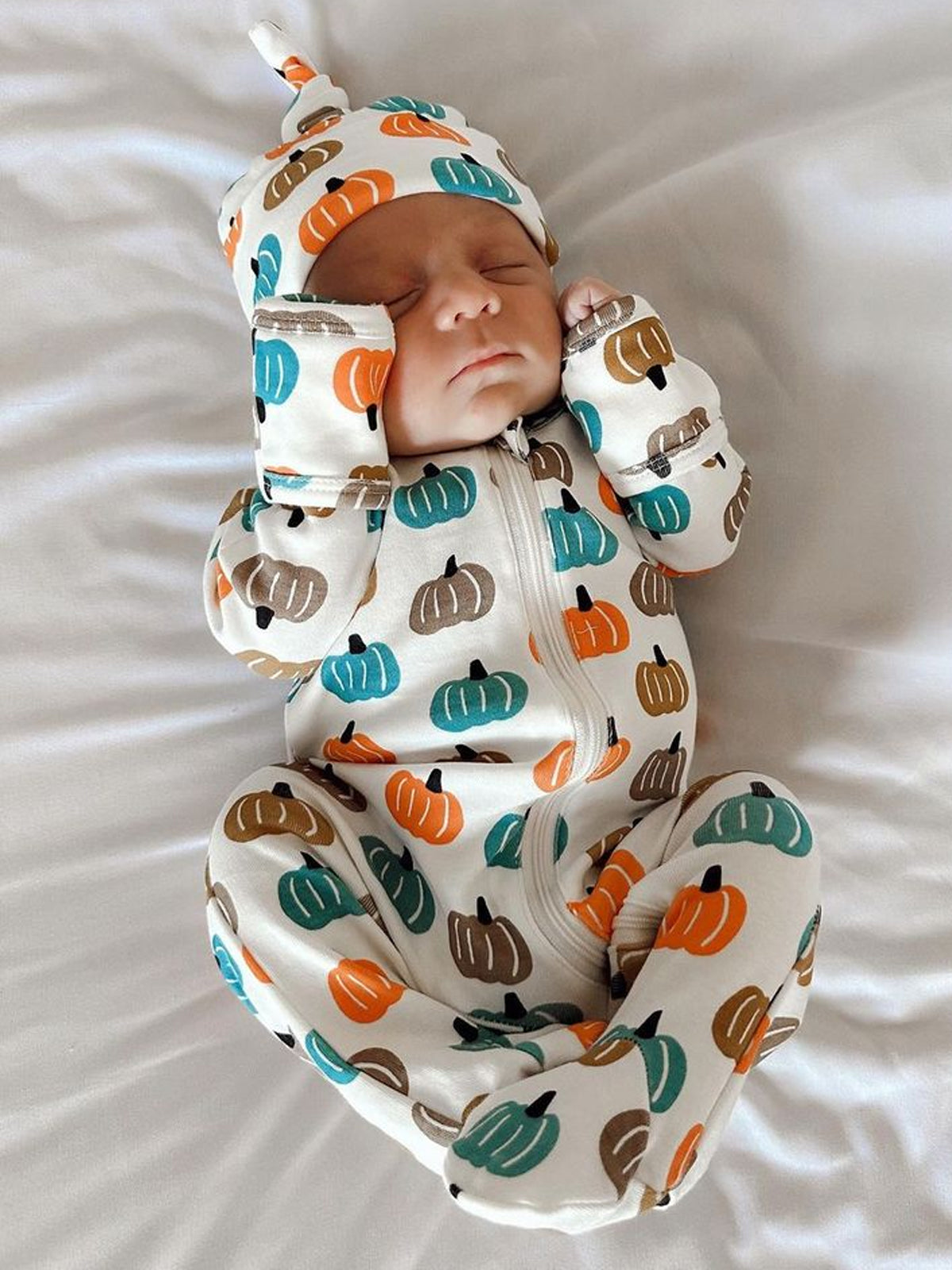 newborn sleeping in pumpkin-patterned outfit on a white blanket
