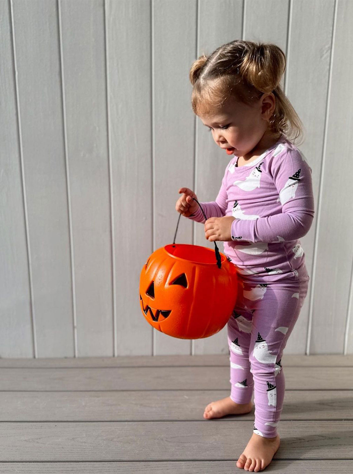 Young child in ghost-patterned pajamas holding a pumpkin candy bucket on a wooden floor, playful Halloween atmosphere.