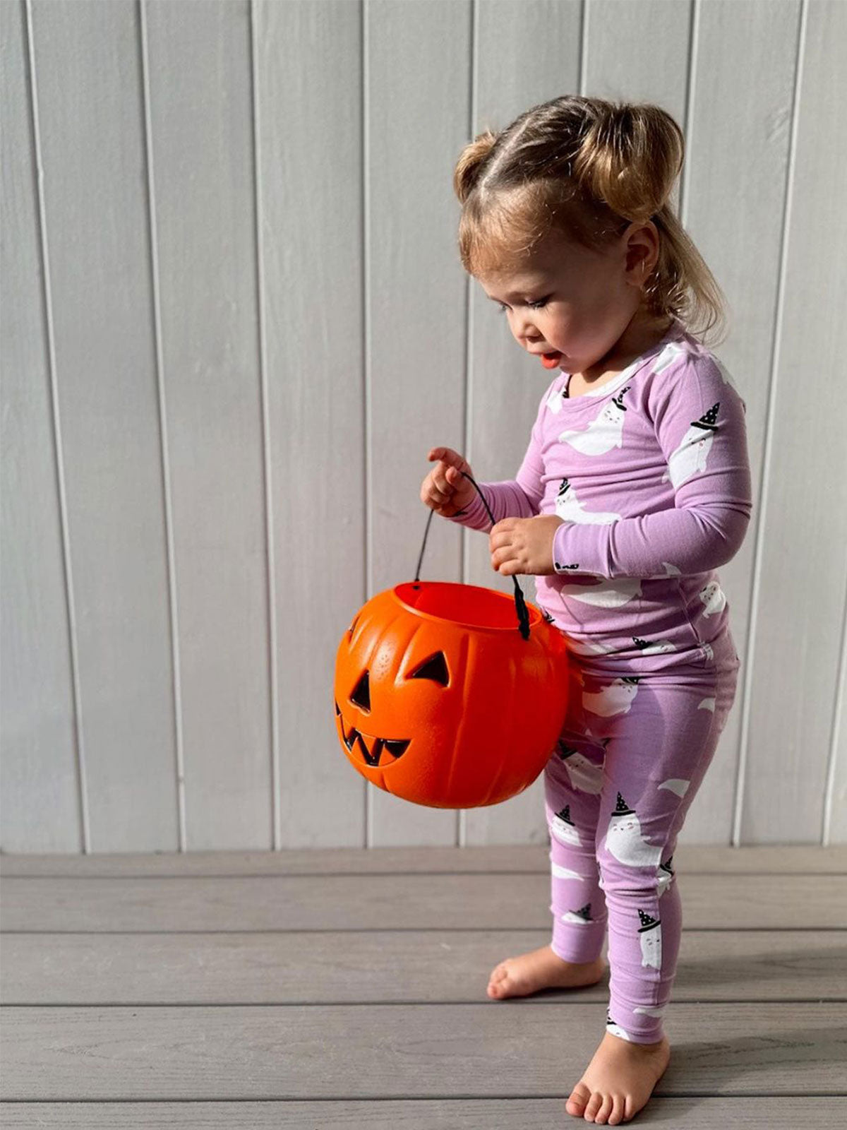 Young child in ghost-patterned pajamas holding a pumpkin candy bucket on a wooden floor, playful Halloween atmosphere.