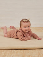 Smiling baby in a pink outfit, lying on stomach on a soft mat, with a light-colored brick wall in the background.