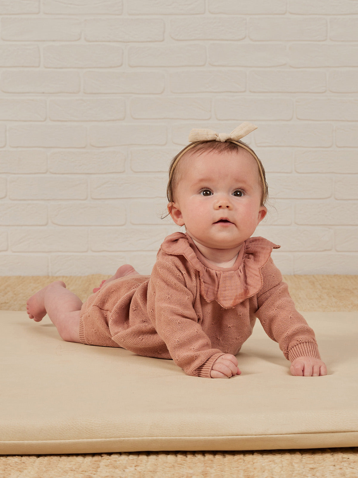 Baby in a cozy pink outfit and bow, crawling on a soft mat against a white brick background.