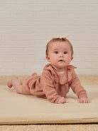 Baby in a cozy pink outfit and bow, crawling on a soft mat against a white brick background.