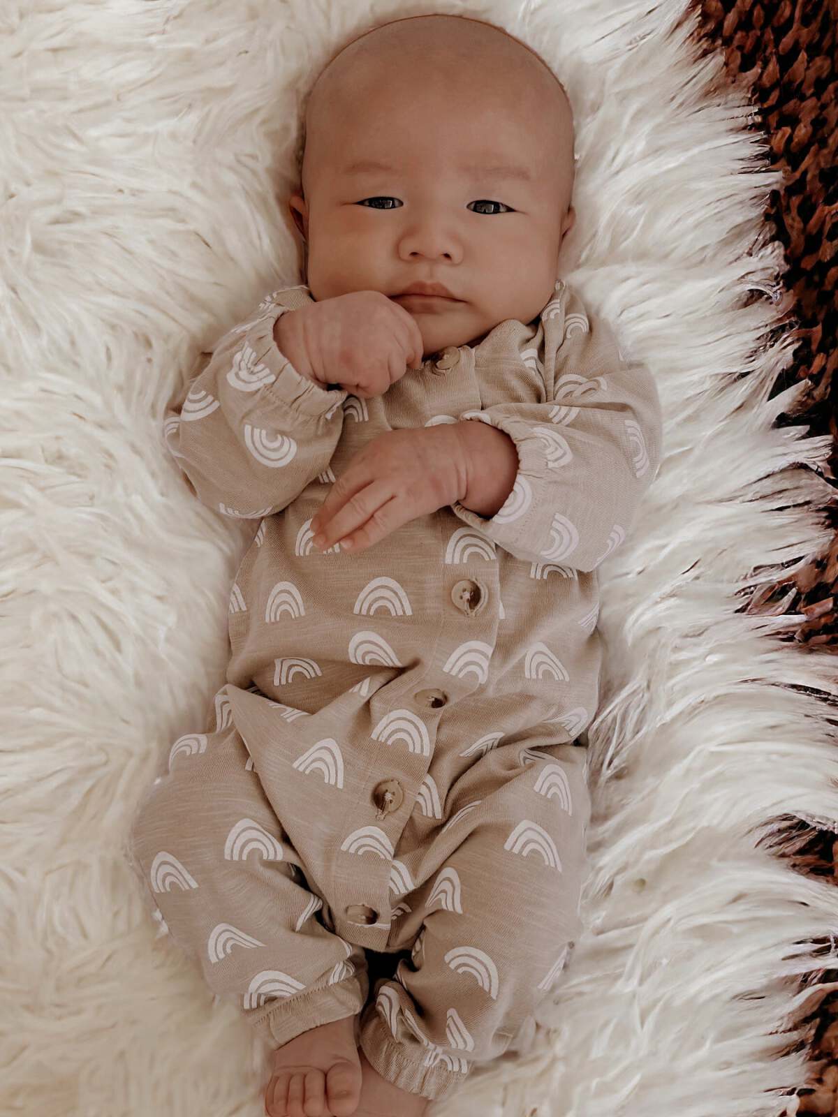 Baby in a cozy rainbow-patterned outfit, lying on a fluffy white rug, looking directly at the camera.