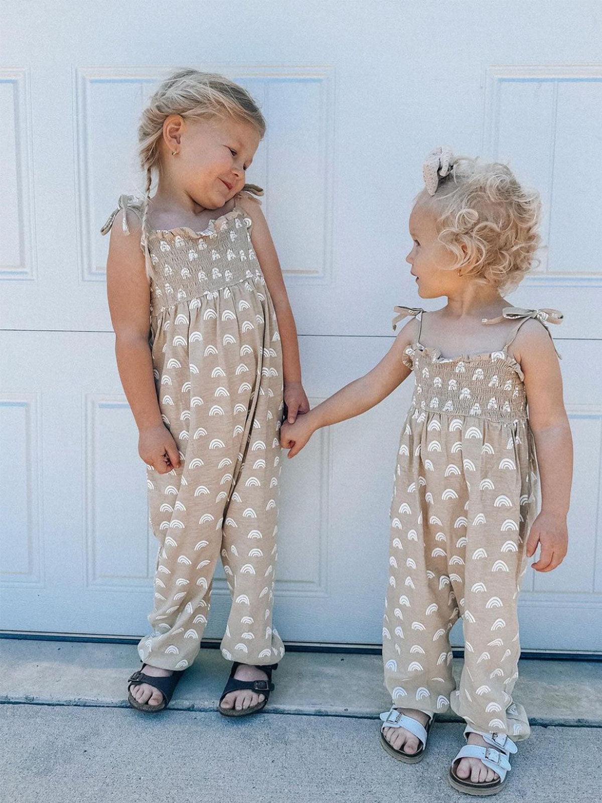 Two young girls in matching tan outfits with white rainbows, holding hands by a garage door.