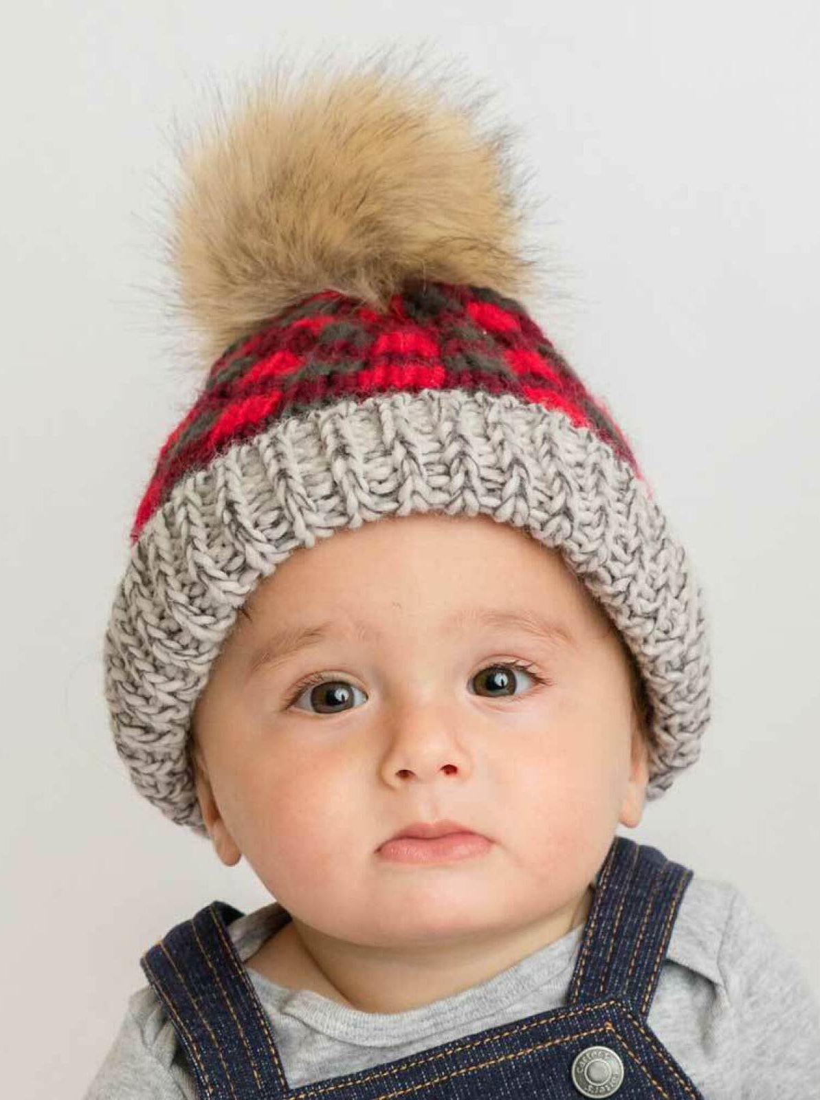 Baby in a knitted hat with a faux fur pom-pom, wearing a gray top and denim overalls, looking at the camera.
