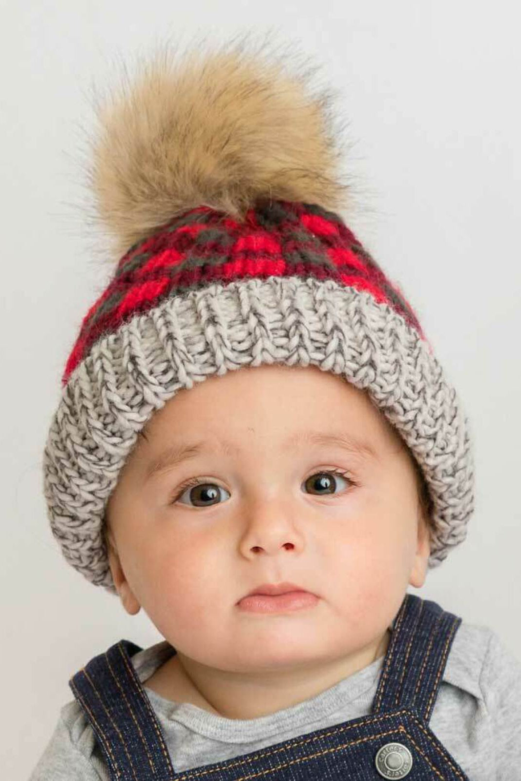 Baby in a knitted hat with a faux fur pom-pom, wearing a gray top and denim overalls, looking at the camera.