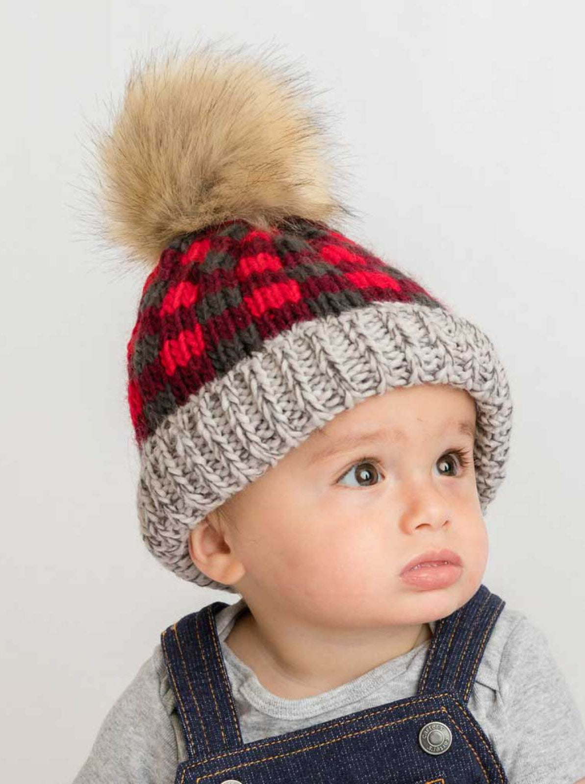 Child wearing a red and gray checkered knit hat with a fur pom-pom. Expression is curious and thoughtful.