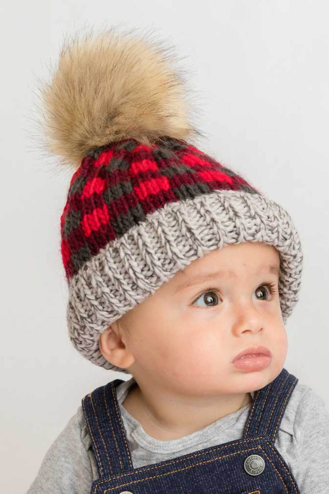 Child wearing a red and gray checkered knit hat with a fur pom-pom. Expression is curious and thoughtful.