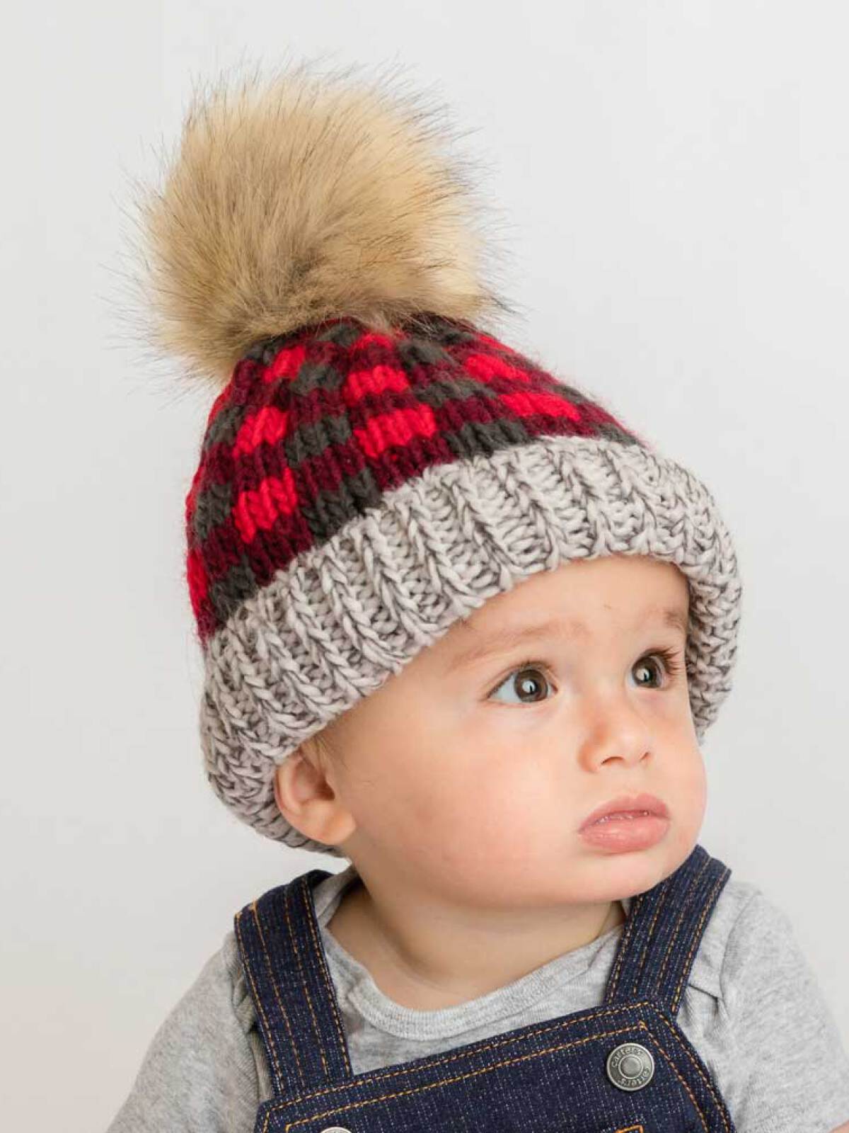 Child wearing a red and gray checkered knit hat with a fur pom-pom. Expression is curious and thoughtful.