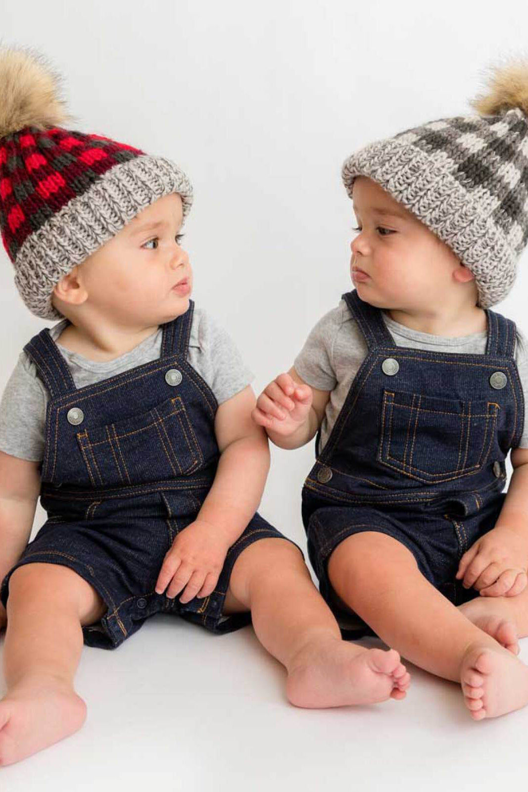 Two babies in denim overalls and knitted hats sit side by side, interacting with each other on a light background.