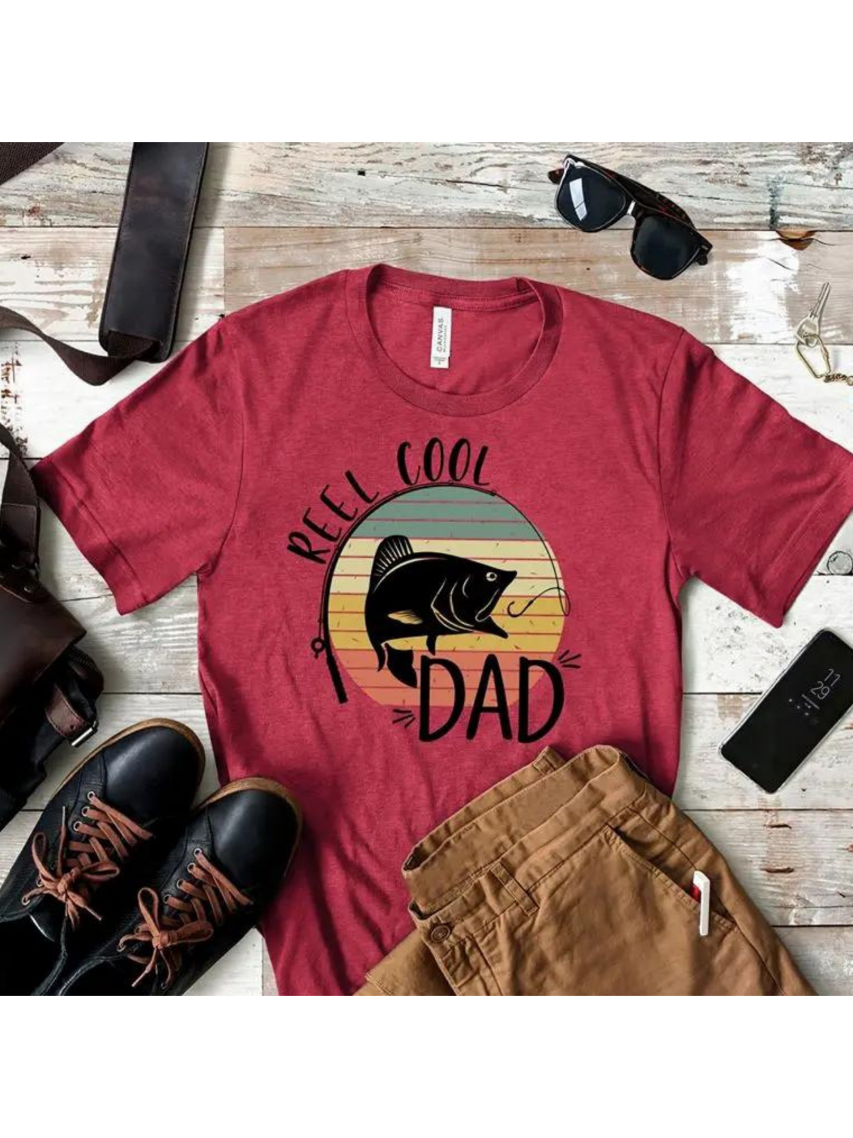 Red t-shirt with "Reel Cool Dad" graphic, surrounded by sunglasses, shoes, and casual pants on a wooden surface.