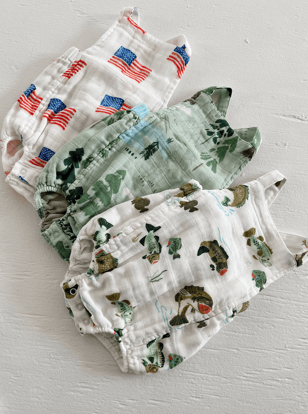 Four colorful infant rompers with patterns: American flags, greenery, and playful fish designs, arranged neatly.