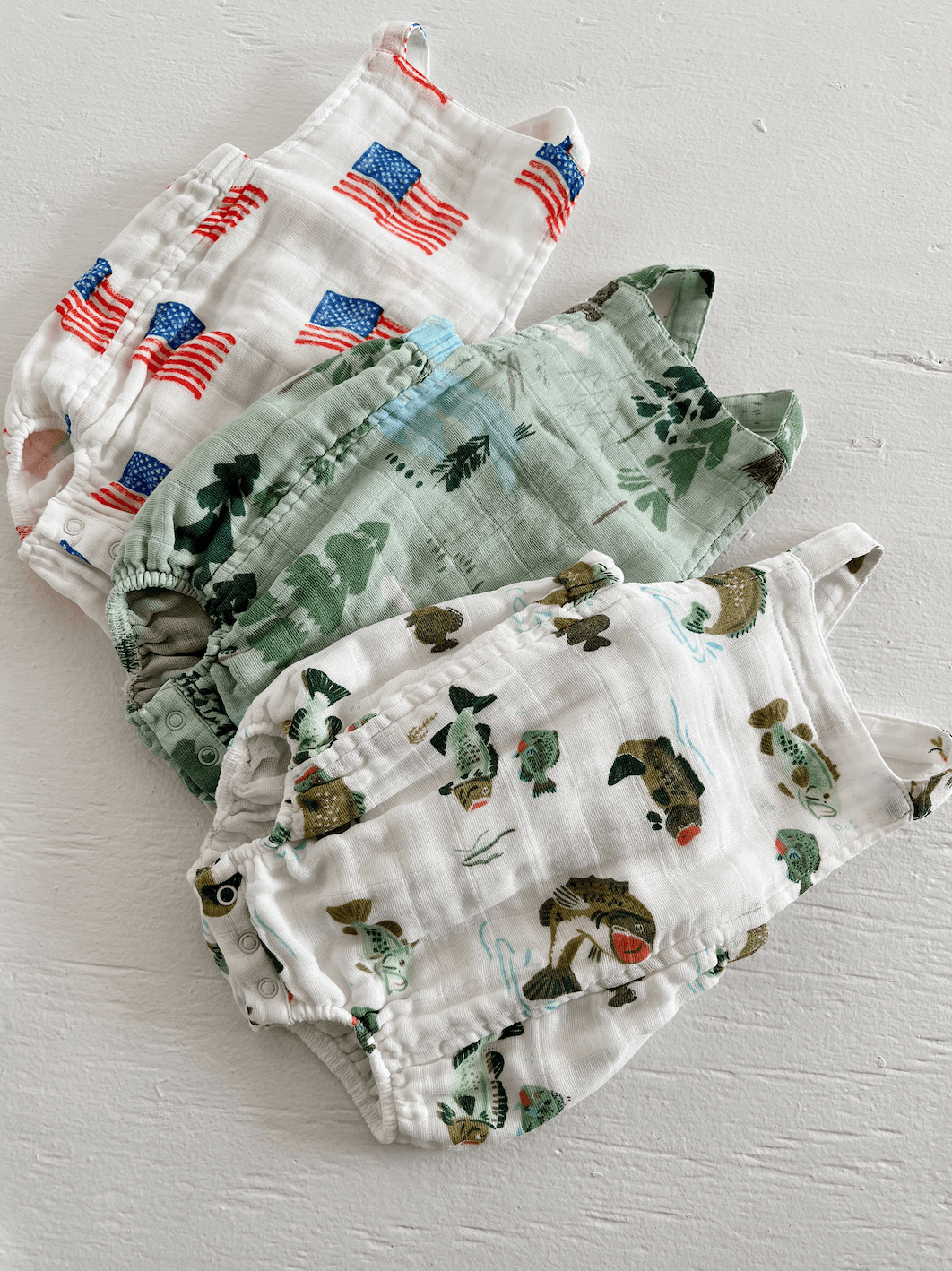 Four colorful infant rompers with patterns: American flags, greenery, and playful fish designs, arranged neatly.