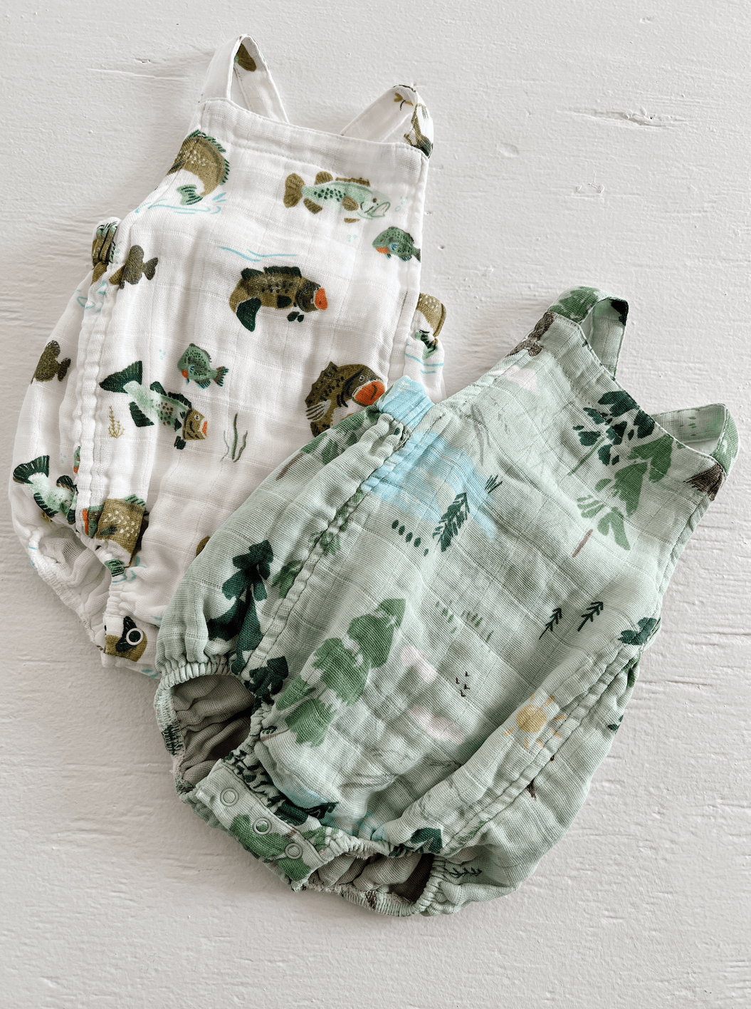 Two baby rompers in a fish pattern and a forest theme on a textured white background.