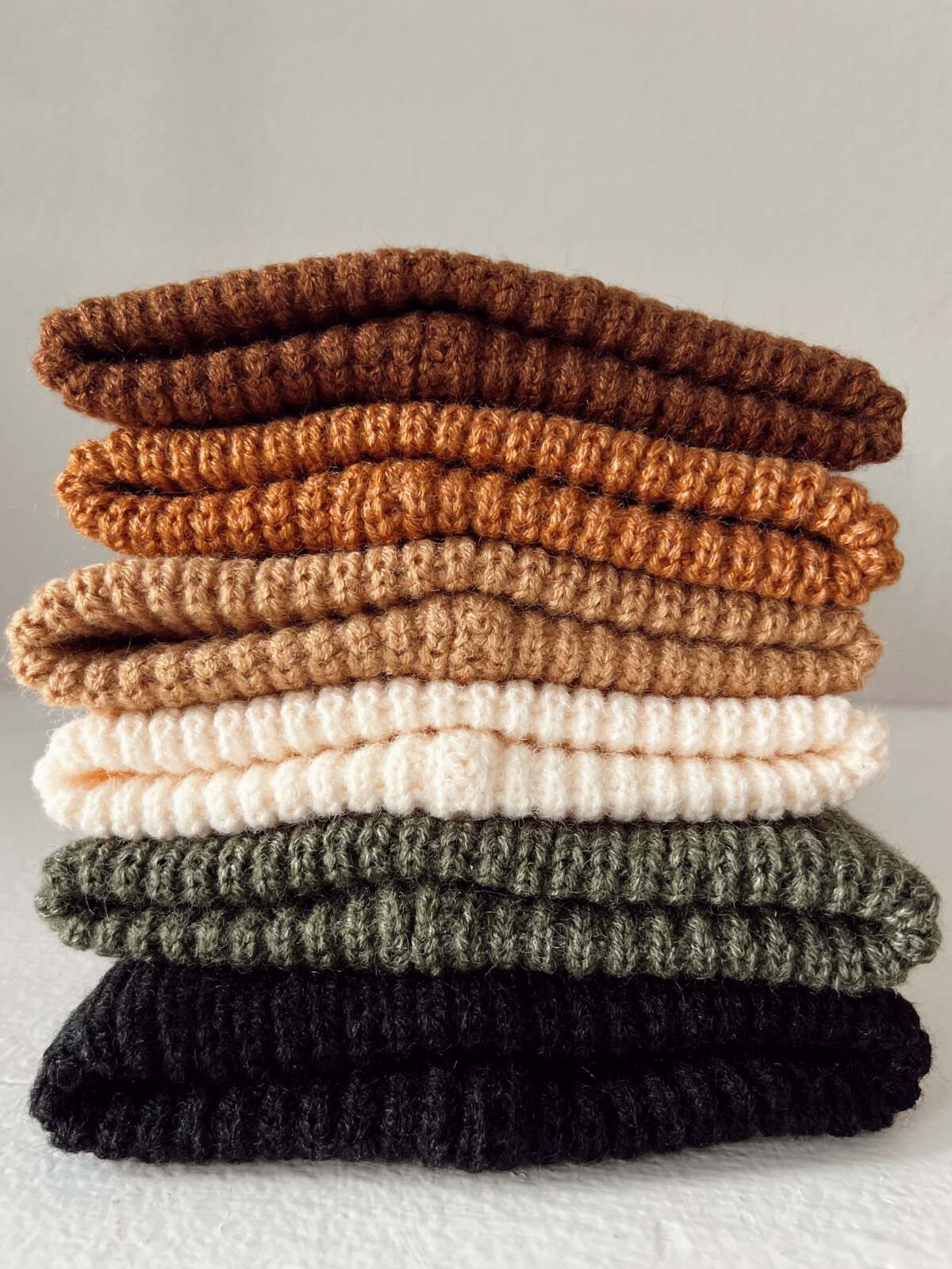 Stack of seven textured knit items in warm earthy colors: brown, tan, cream, green, and black, against a neutral background.