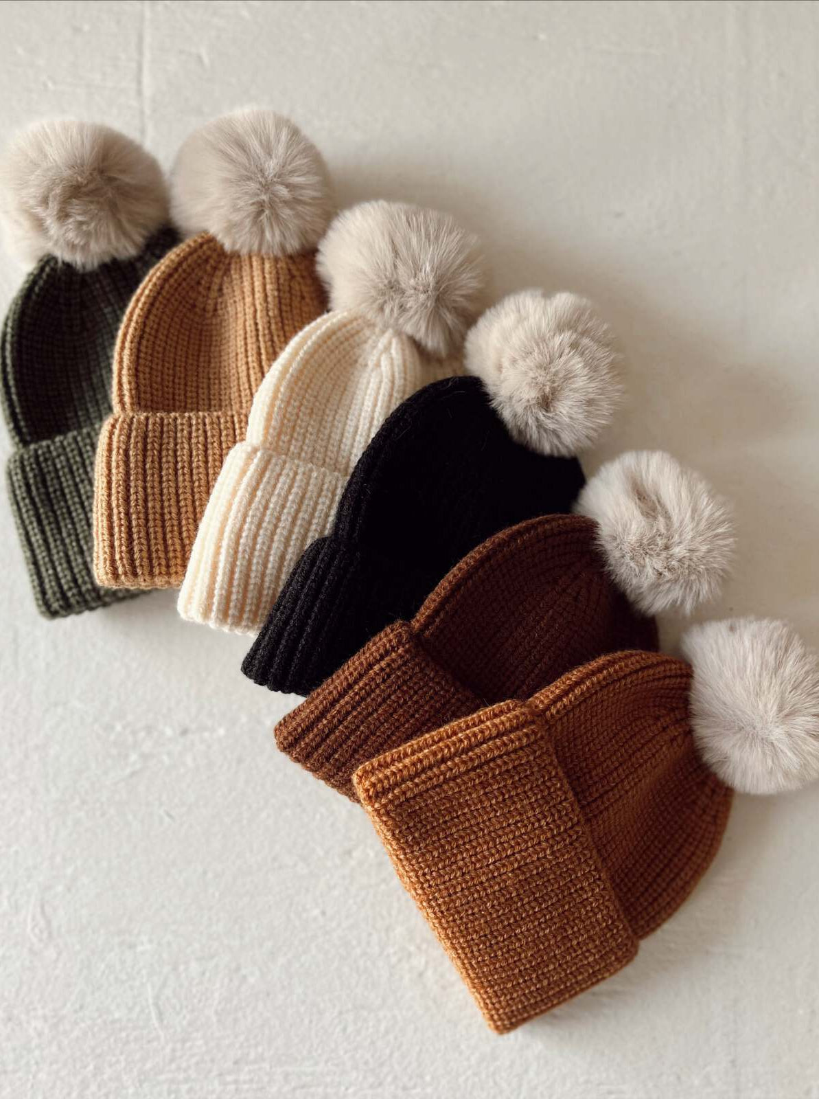 A selection of six knitted beanies with pom poms in green, tan, white, black, brown, and rust colors.