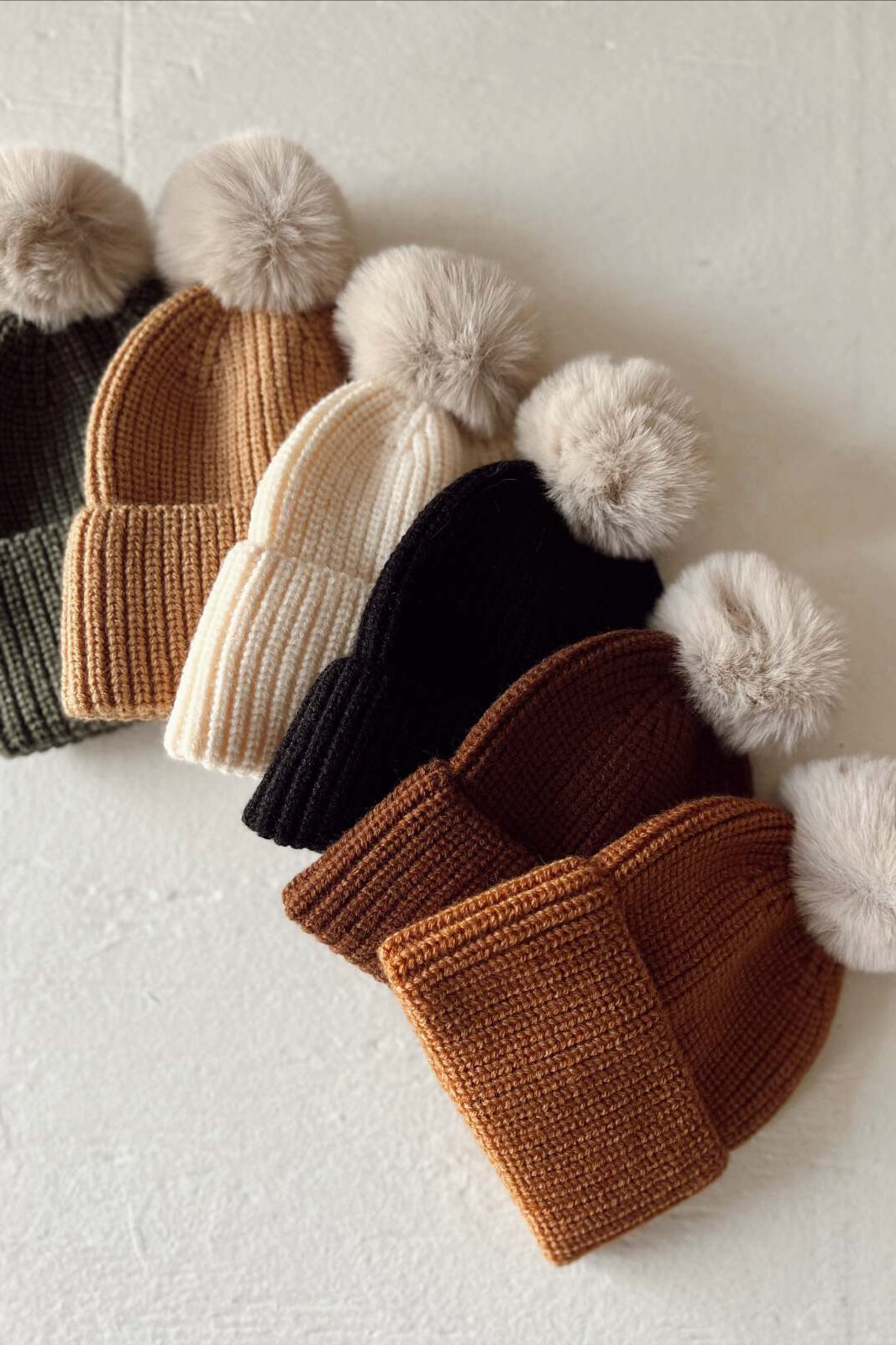 A collection of five cozy knit beanies in green, tan, cream, black, and brown, each featuring a fluffy pom-pom.